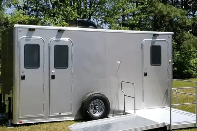 A silver shower restroom trailer with three doors, a spare tire, and an attached wheelchair-accessible ramp is parked outdoors near trees.