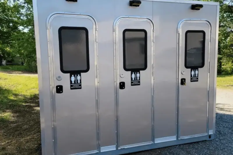 A portable shower restroom trailer with three individual doors, each labeled for both male and female use, is situated outdoors on a paved surface.
