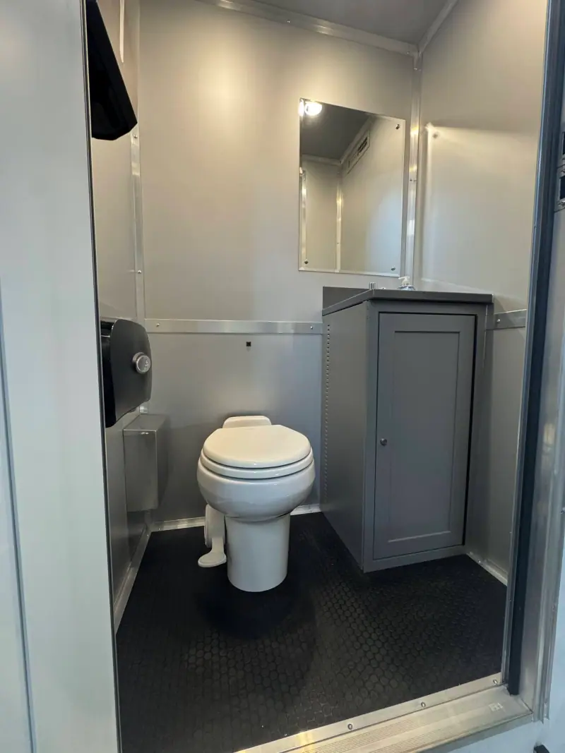 The ADA+4 restroom trailer features a clean, modern design with a white toilet, black toilet paper dispenser, gray cabinet, wall mirror, and metallic walls.
