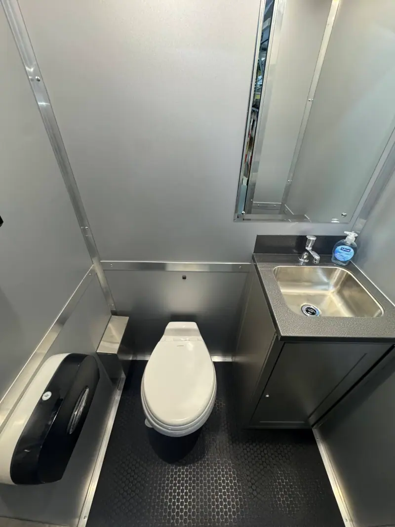The ADA+4 Restroom Trailer features a small, clean space with a white toilet, stainless steel sink, soap dispenser, large mirror, and a toilet paper dispenser.