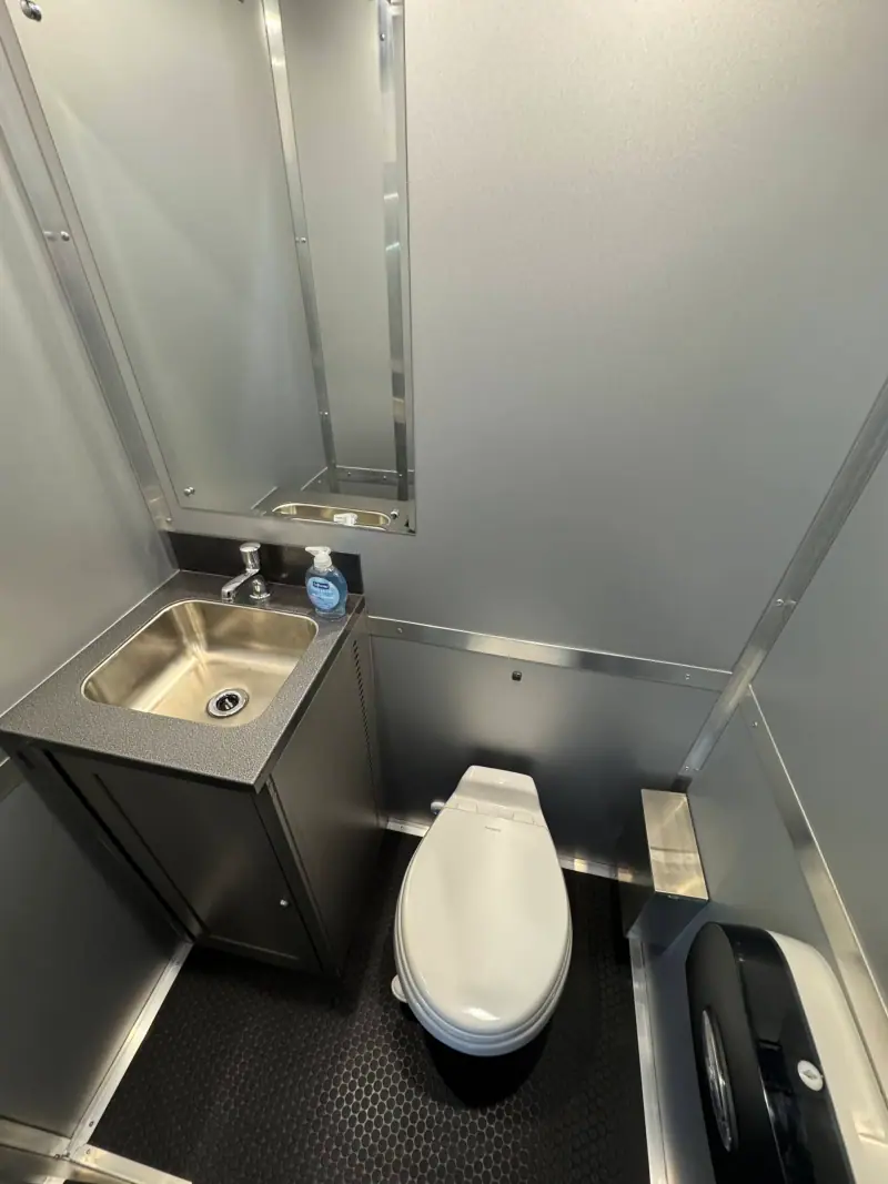 The ADA+4 Restroom Trailer features a compact design with metallic walls, black flooring, a stainless steel sink, soap dispenser, mirror, toilet, and toilet paper dispenser.