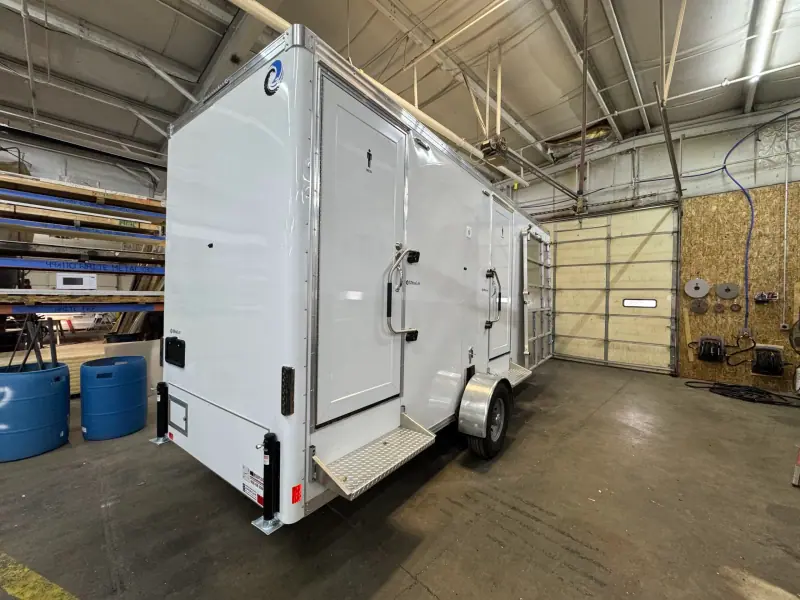 An ADA+4 restroom trailer, featuring white exterior, steps, and handrails, is parked inside a spacious industrial garage with high ceilings and exposed beams.