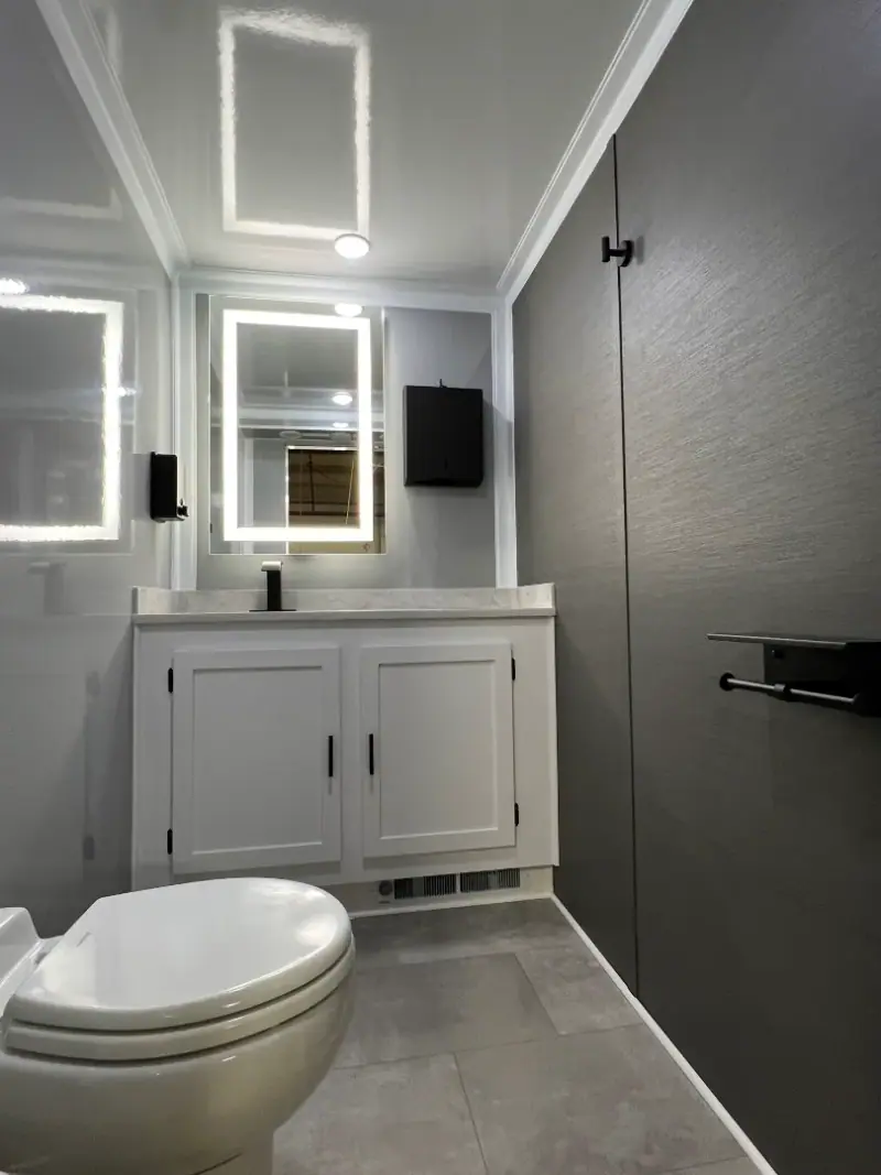 Modern bathroom inside an OI-212 2 Person Stall Restroom Trailer (12' x 6') featuring gray walls, white vanity cabinets, a backlit mirror, wall-mounted soap dispenser, and a toilet in the foreground.