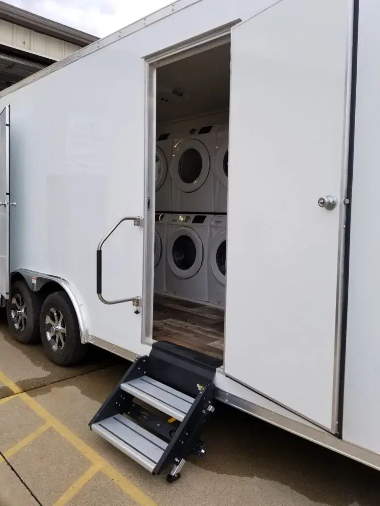 A Laundry Trailer with an open door reveals stacked washers and dryers inside, accessible via a small set of steps.
