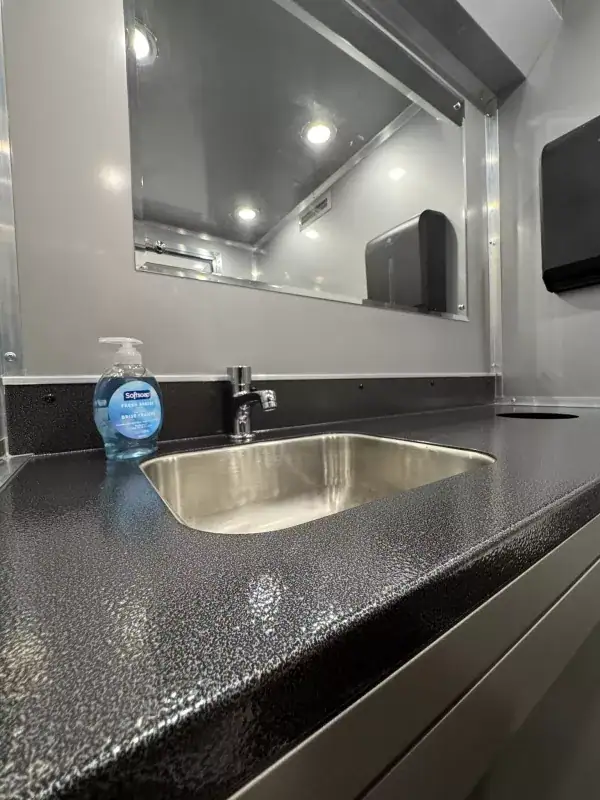 The 3 private room restroom trailer features a stainless steel sink with a faucet on a black countertop, alongside a soap dispenser and beneath a large mirror in its modern interior.