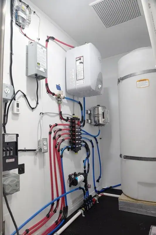 A 3 private room restroom trailer features a utility area with a water heater, large storage tank, electrical panels, and neatly arranged red and blue water pipes set against white walls.