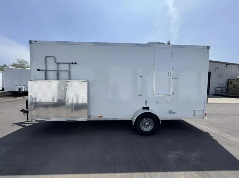 An ADA+2 Restroom Trailer (20' x 6') with a single axle is parked on pavement by an industrial building, featuring visible pipes and a metal box mounted on its side.