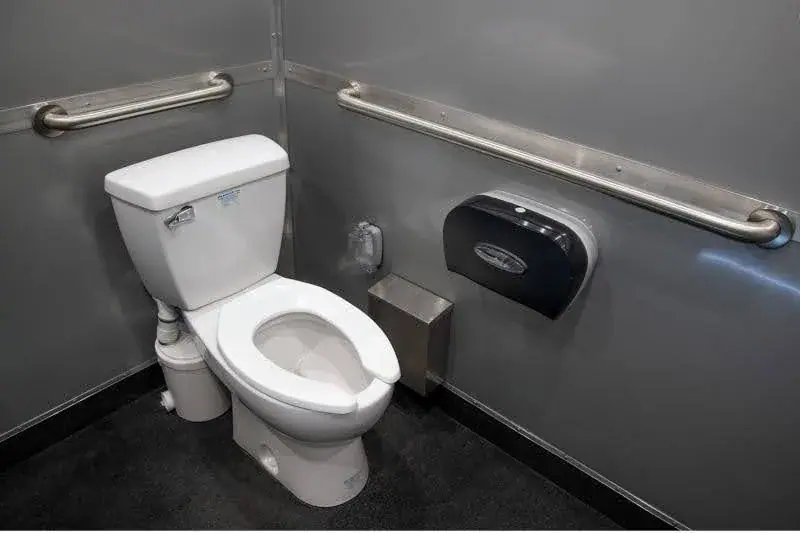 An ADA Single Restroom and Restroom Shower Trailer features a toilet, toilet paper dispenser, trash bin, and two metal grab bars installed on gray walls.