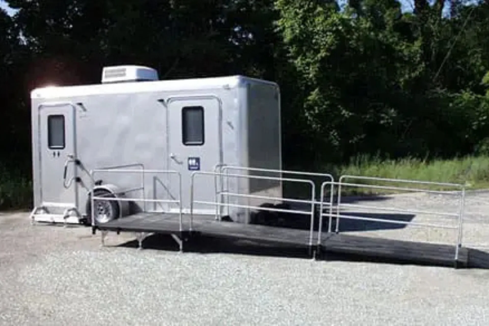 A shower restroom trailer with an accessible ramp is parked on a gravel surface near trees and grass.
