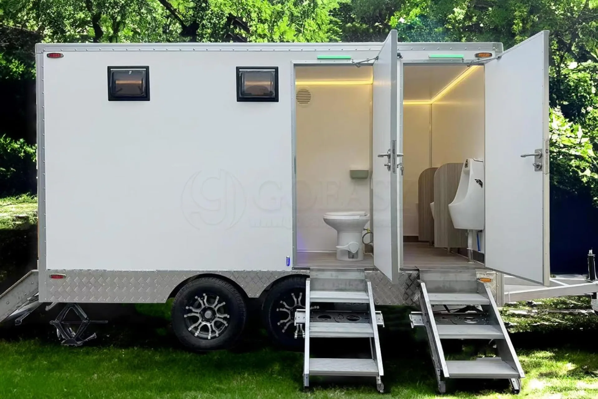 A portable shower restroom trailer with open doors reveals a toilet, urinal, and steps leading up to the entrance, set outdoors near greenery.