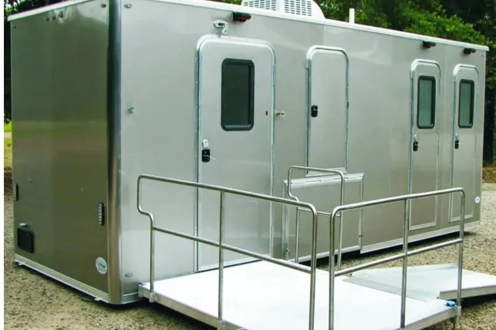 A portable shower restroom trailer with three separate doors and windows, featuring a wheelchair accessible ramp in front, is situated outdoors on gravel.