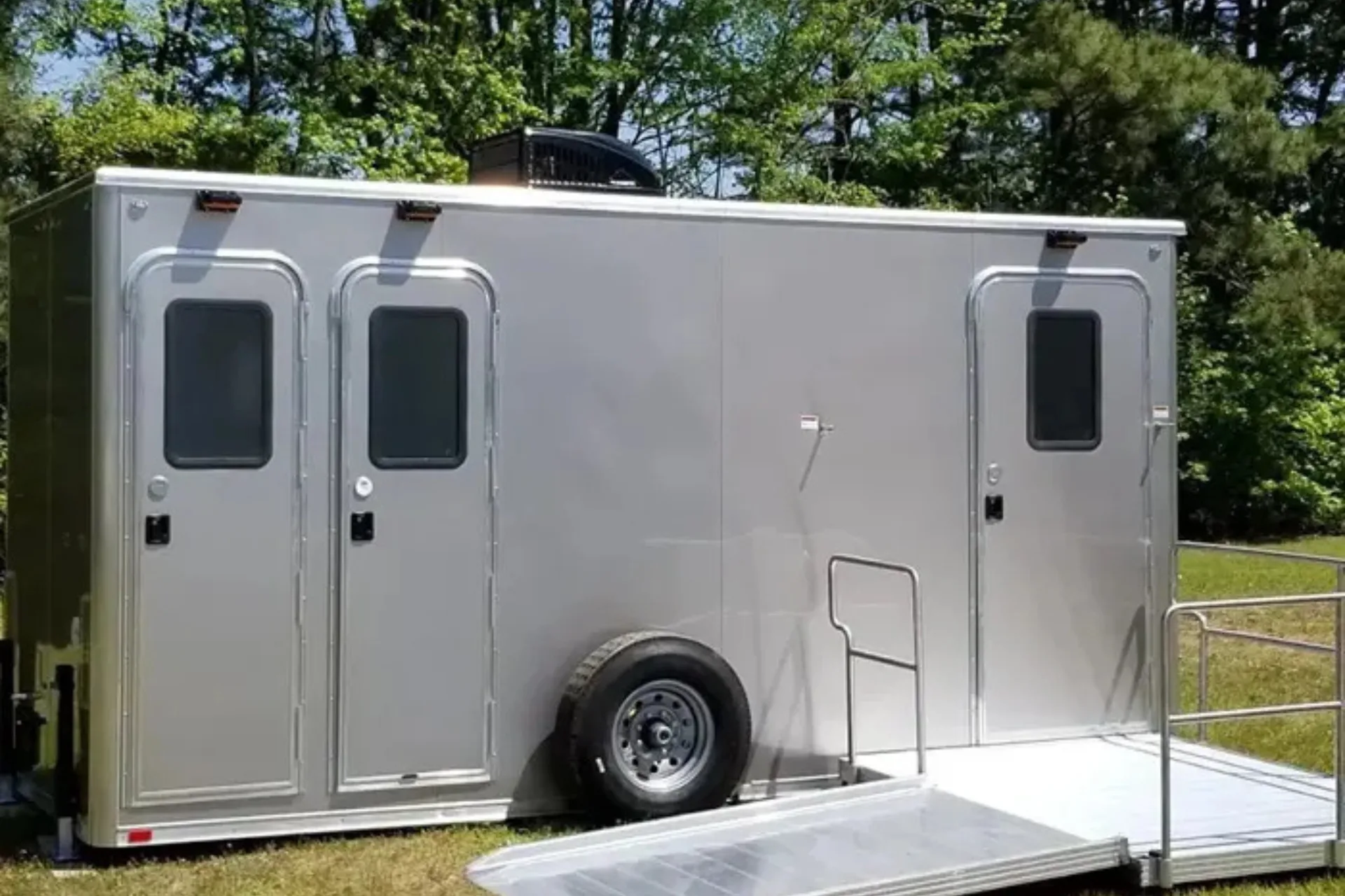 A silver shower restroom trailer with three doors, a spare tire, and an attached wheelchair-accessible ramp is parked outdoors near trees.
