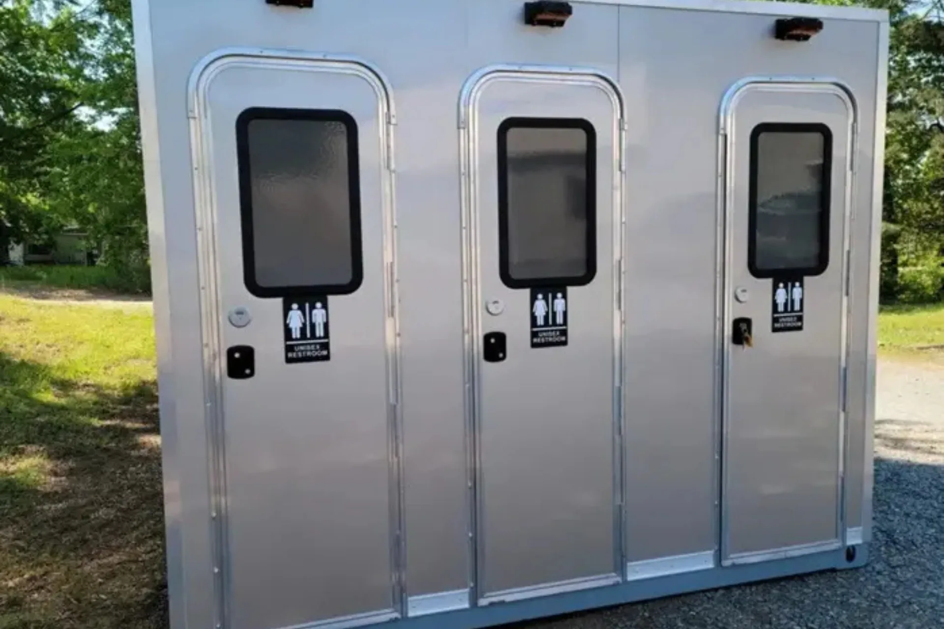 A portable shower restroom trailer with three individual doors, each labeled for both male and female use, is situated outdoors on a paved surface.