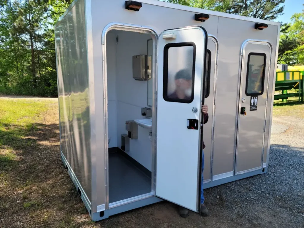 A person is exiting a shower restroom trailer with multiple stalls, situated outdoors on a gravel and grass surface.