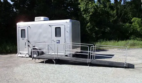A silver shower restroom trailer with a wheelchair-accessible ramp is parked on a gravel lot near trees.
