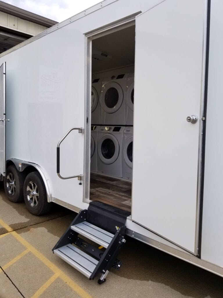 A Laundry Trailer with an open door reveals stacked washers and dryers inside, accessible via a small set of steps.