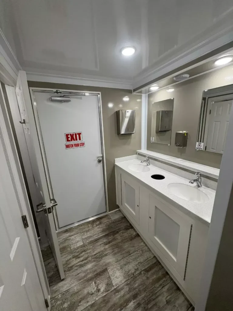 A clean shower restroom trailer with two sinks, mirrors, a soap dispenser, and an EXIT door featuring a sign that reads "EXIT WATCH YOUR STEP" on the back wall.
