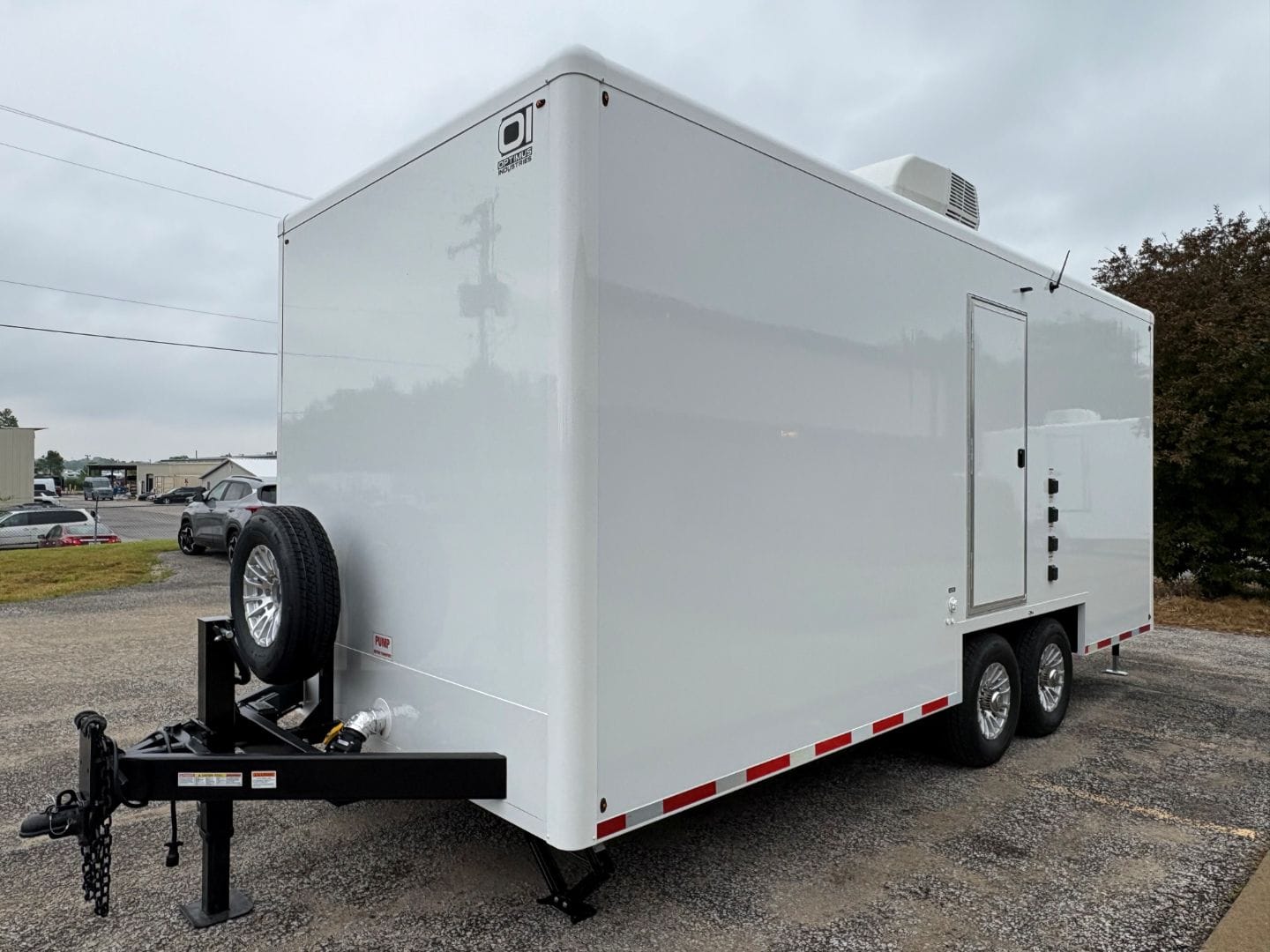 The OI-620 - 6 Station Restroom Trailer (20' x 8.5') is a white dual-axle unit with a side door, rooftop vent, and mounted spare tire, shown parked on gravel with other vehicles and trees in the background.