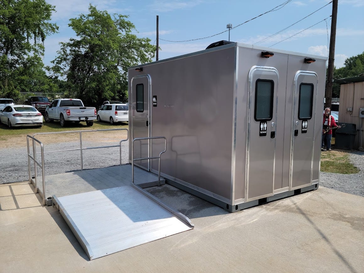 A CI 12 - 3 Stall ADA Restroom Skid (12 ft) with a metal wheelchair ramp is set up on a concrete surface in an outdoor parking lot.