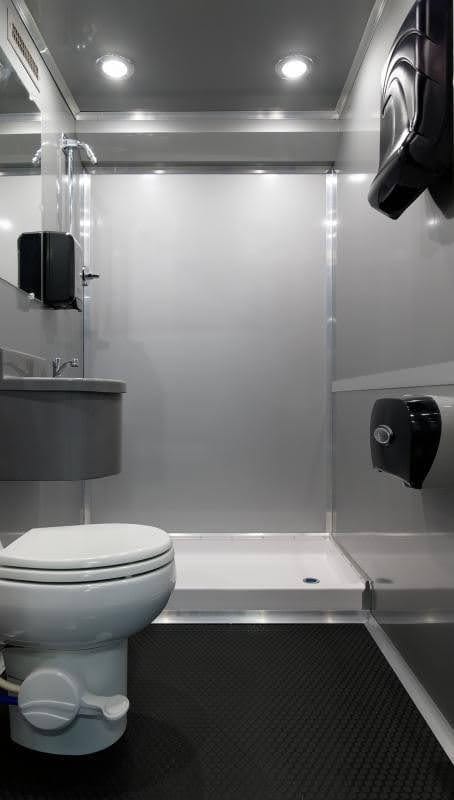 The 2 private room shower restroom trailer features a modern, compact bathroom with a white toilet, shower area, wall-mounted sink, mirror, soap and toilet paper dispensers on gray walls with rubber flooring.