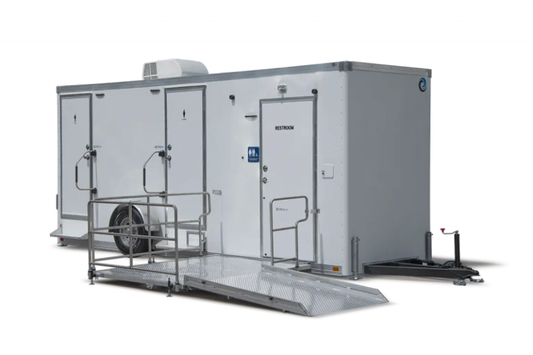 White portable restroom trailer with three doors, accessibility ramp, and metal railings, set up for temporary or outdoor use.