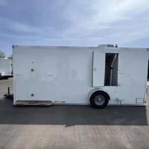 A plain white enclosed shower restroom trailer with one open side door is parked on a paved lot under a partly cloudy sky.