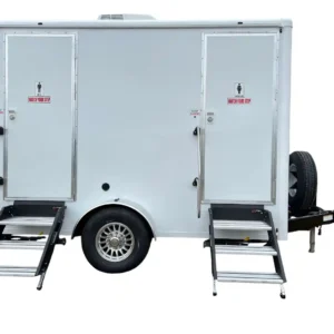 The OI-212 — 2 Person Stall Restroom Trailer (12' x 6') is a white mobile unit with two doors, metal step ladders for access, and a spare tire mounted on the front.
