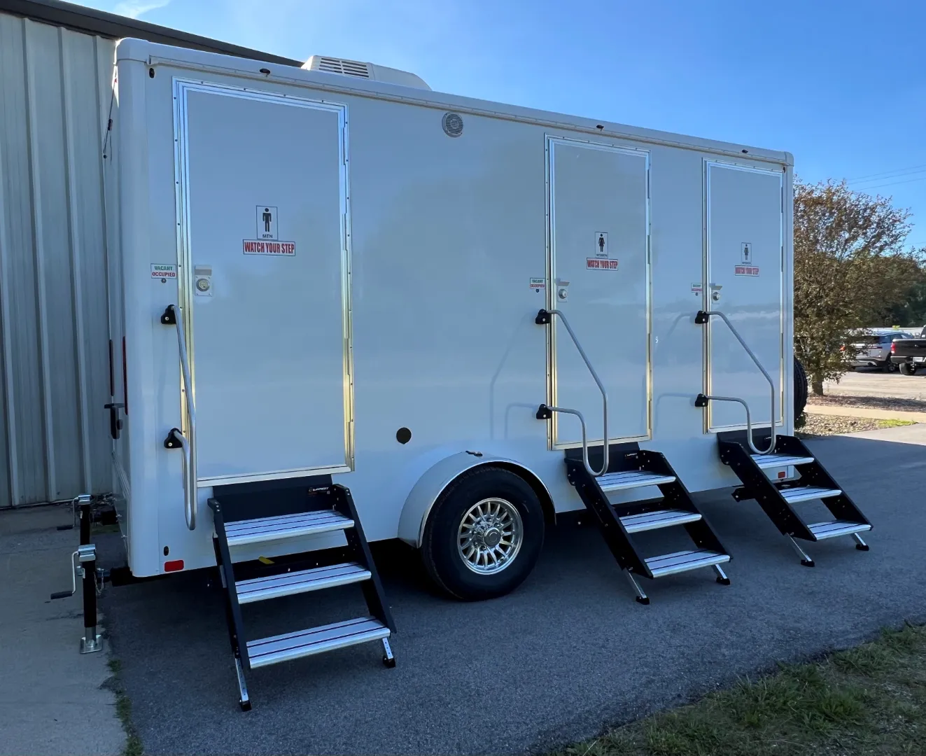 A white mobile restroom trailer with three separate entry doors, each with stairs leading up to the door, is parked next to a building.