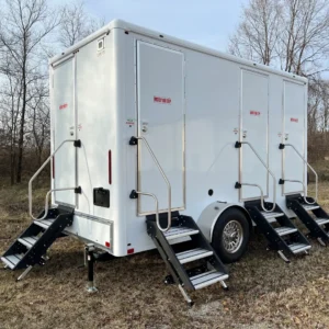 A portable shower restroom trailer with three separate doors and staircases stands on a grassy area, with leafless trees in the background.