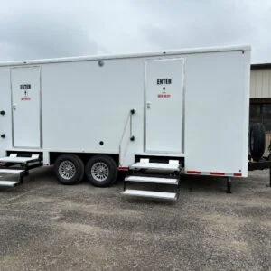 A white shower restroom trailer with two doors marked “Enter” stands on gravel, each door accessed by metal steps.
