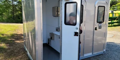 A person is exiting a shower restroom trailer with multiple stalls, situated outdoors on a gravel and grass surface.