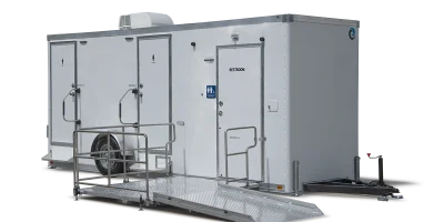 A portable restroom trailer with three doors, including accessible entry with a ramp, air conditioning unit on top, and a white exterior.