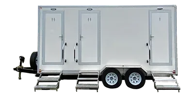 A white shower restroom trailer with three doors, each with a step leading up, mounted on a double-axle trailer.